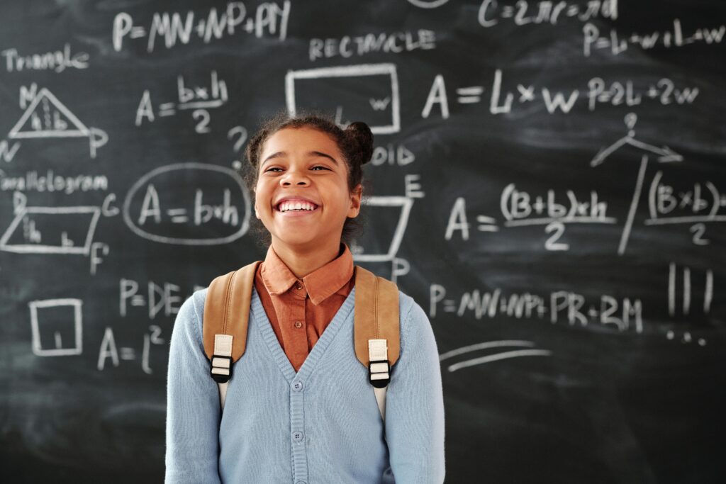 Smiling girl with backpack in a classroom, standing in front of a chalkboard with math formulas.