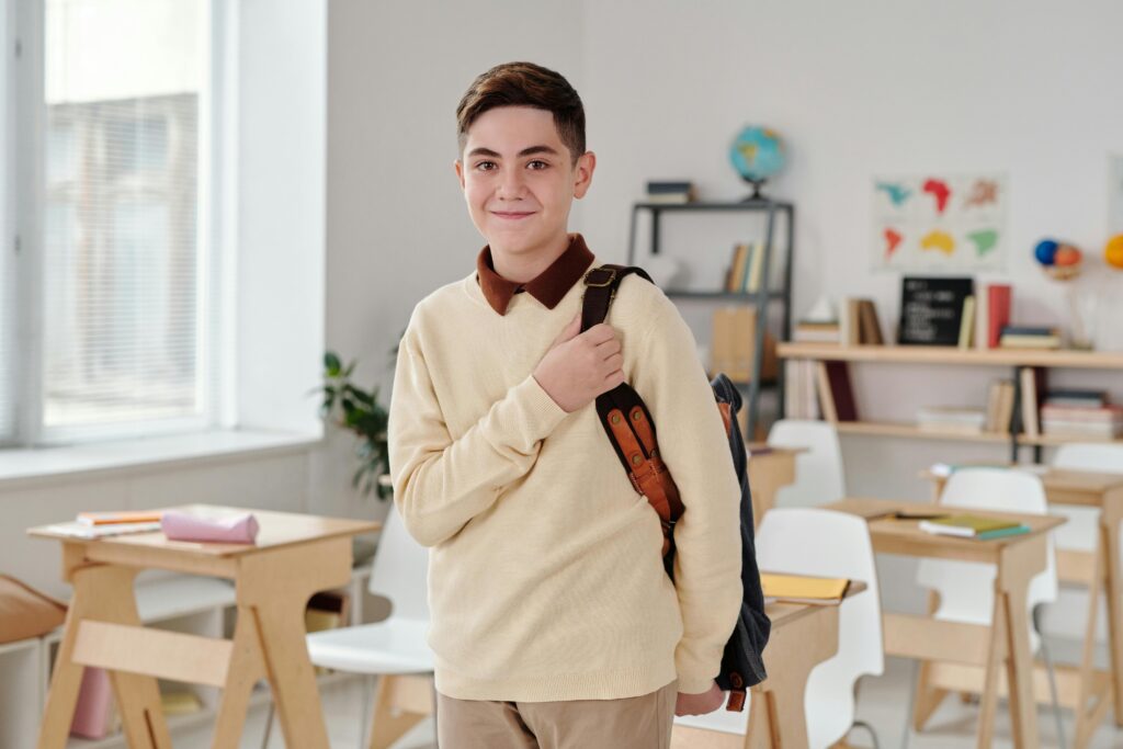 A smiling schoolboy confidently standing in a modern classroom with his backpack.